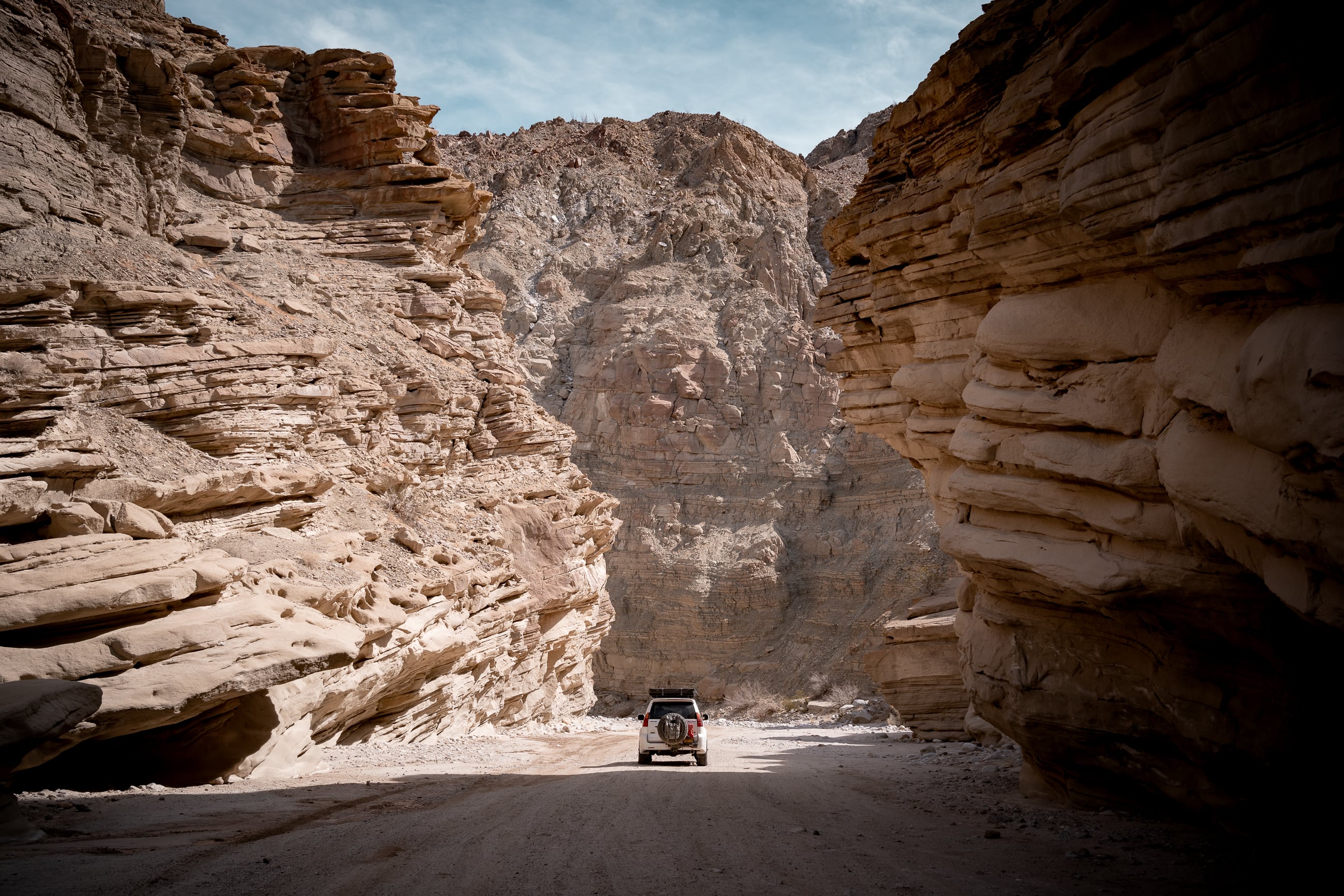 A single SUV, a LexusGX470 sits in the middle of a canyon, dwarfed by sandy brown craggy layers of rock walls.
